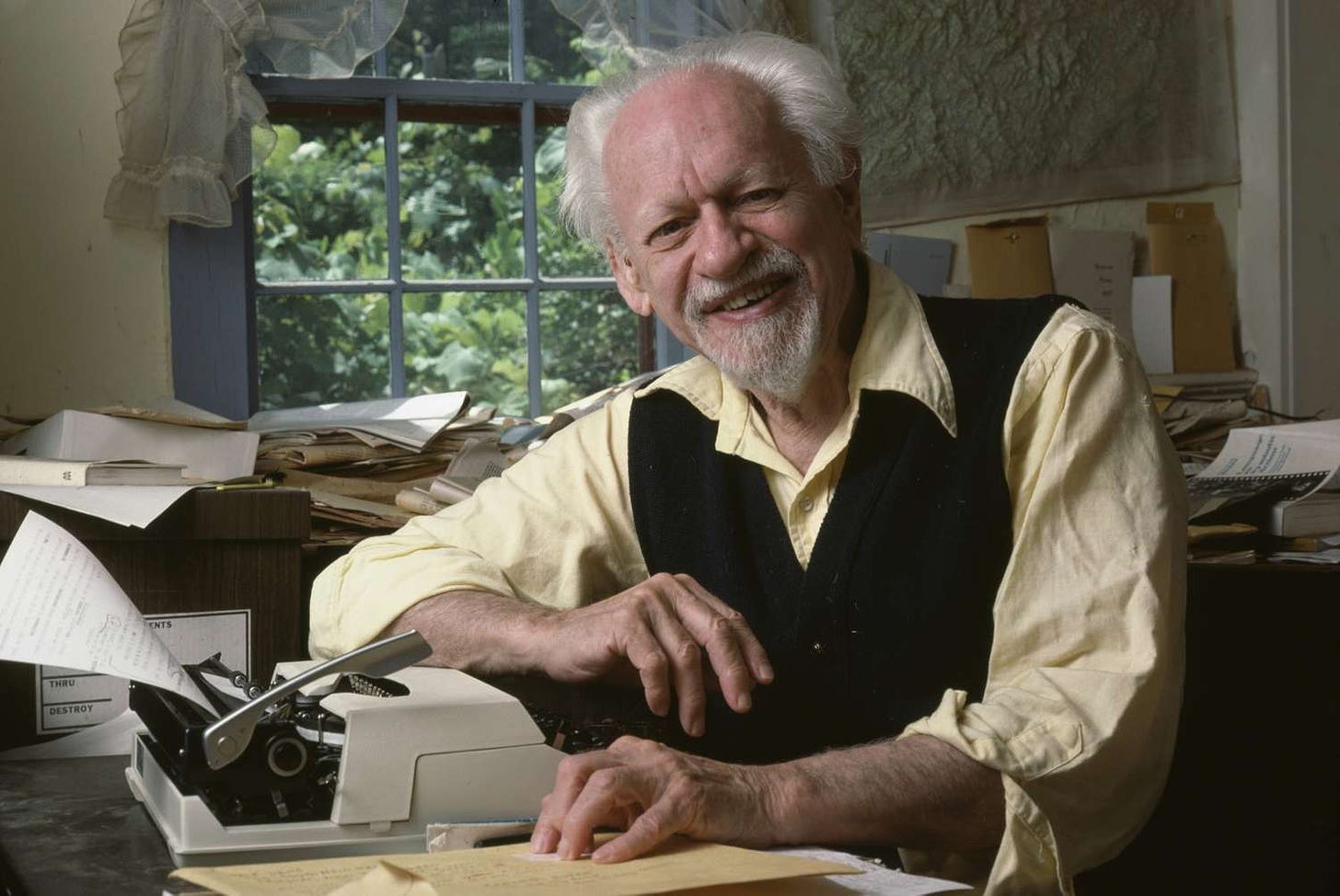 Kenneth Burke, an old man with white hair and a goatee, smiles somewhat awkwardly at the camera while sitting in front of a typewriter.