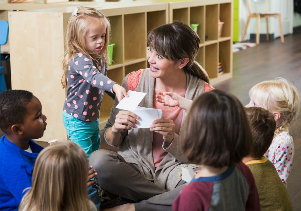 Female primary school teacher sitting on floor with infants and girl with learning disability helping to hand out plain white cards
