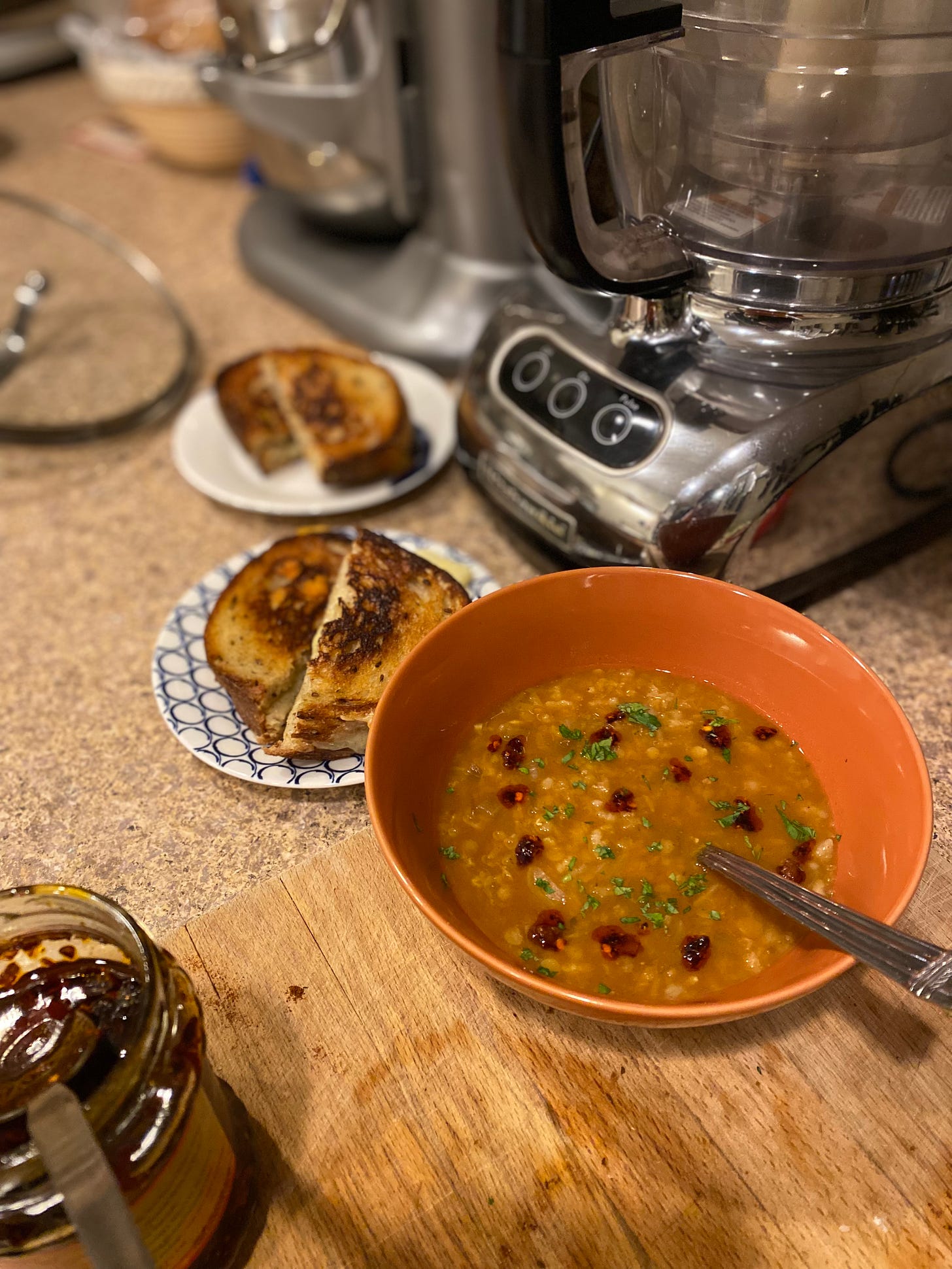 An bright orange bowl of yellow-orange soup dotted with chili crisp and parsley, a spoon sticking out of it. Behind it on the counter are two small plates, each with a grilled cheese sliced in half. In the foreground, the jar of chili crisp is visible with a small spoon in it.