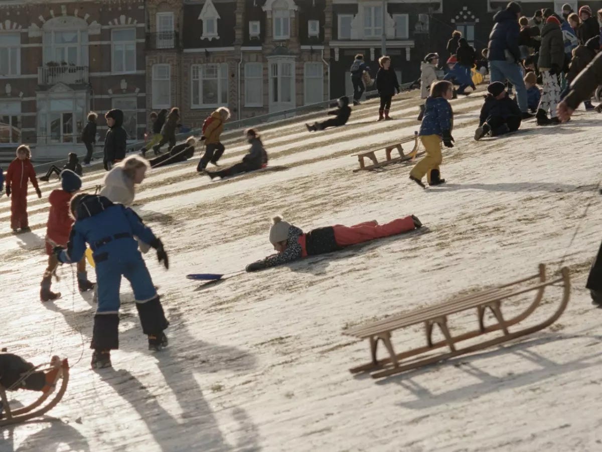 A snowy slope with many wooden sleds and children in snow suits. One child in an orange suit and pom pom hat is lying stomach-down in the snow. Behind the scene we see a row of brick houses in the Amsterdam style. 