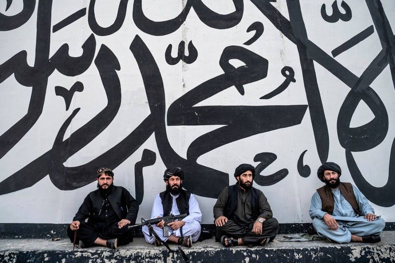 Four men with rifles sit on the ground against a wall painted with Arabic script.