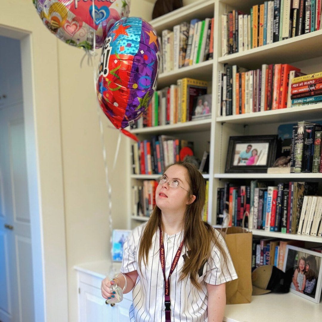 Penny stands in a room filled with bookshelves. She is wearing a white and black striped shirt and a lanyard around her neck, looking up at two colorful balloons she's holding by the strings. One balloon has vibrant stars and patterns, while the other has "Congrats" written on it. Behind her, the shelves are lined with books and family photos, giving the space a warm, personal feel