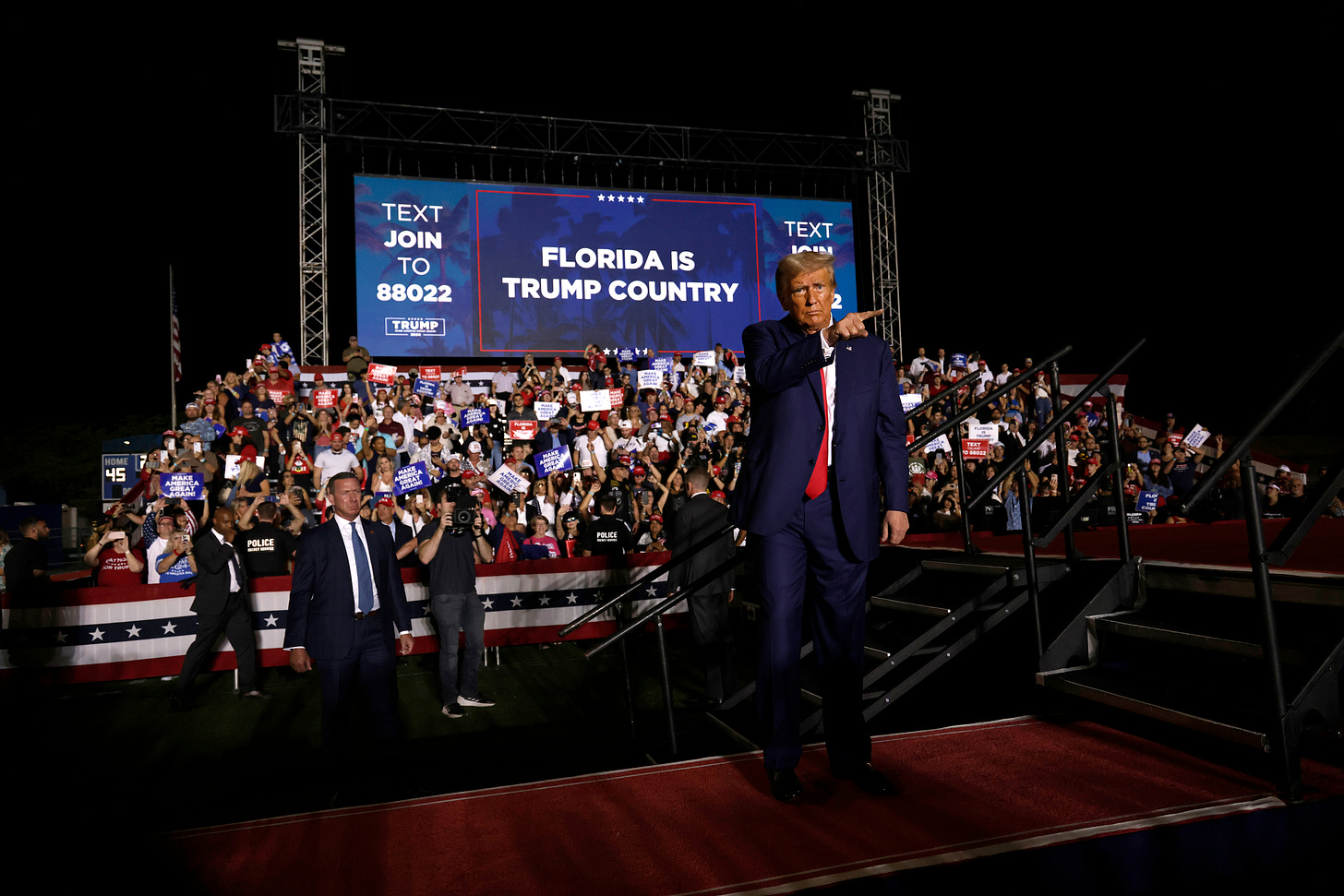 A blonde man in a dark suit stands in front of a screen that reads "Florida is Trump Country"