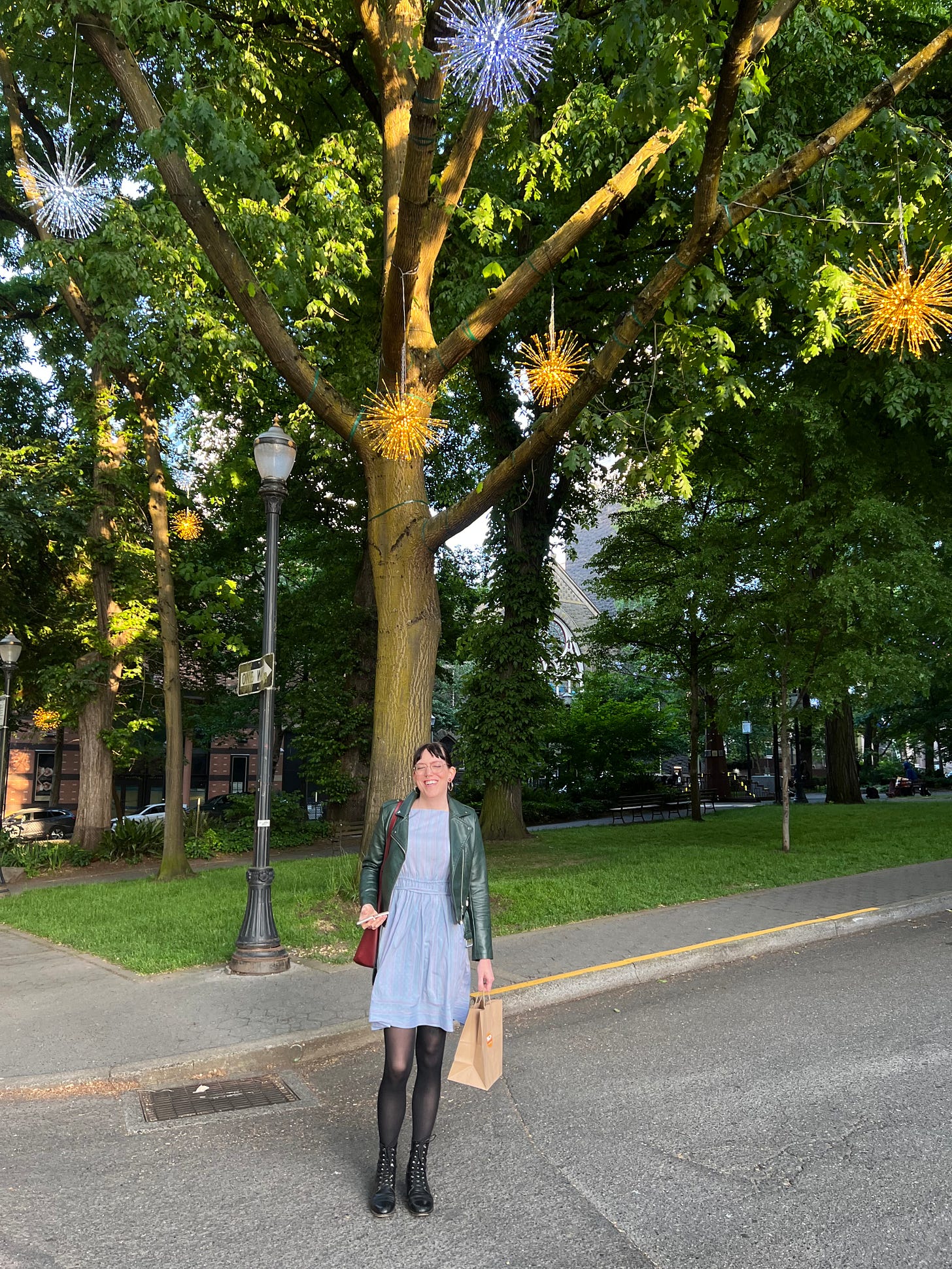 Emme stands in a blue dress and tights under a tree in the park blocks in downtown portland. Twinkling light globes hang in the tree above her. The light is golden.