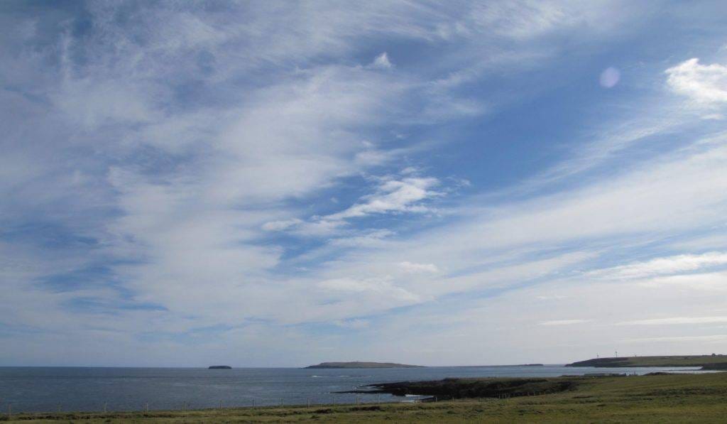A view of an island in the east of Orkney, now uninhabited. The sky is blue but there are clouds, the sea relatively calm. These islands are ancient, the landscape inspiring and creating legends, art, stories, poetry, and tales of travel and adventure for thousands of years.