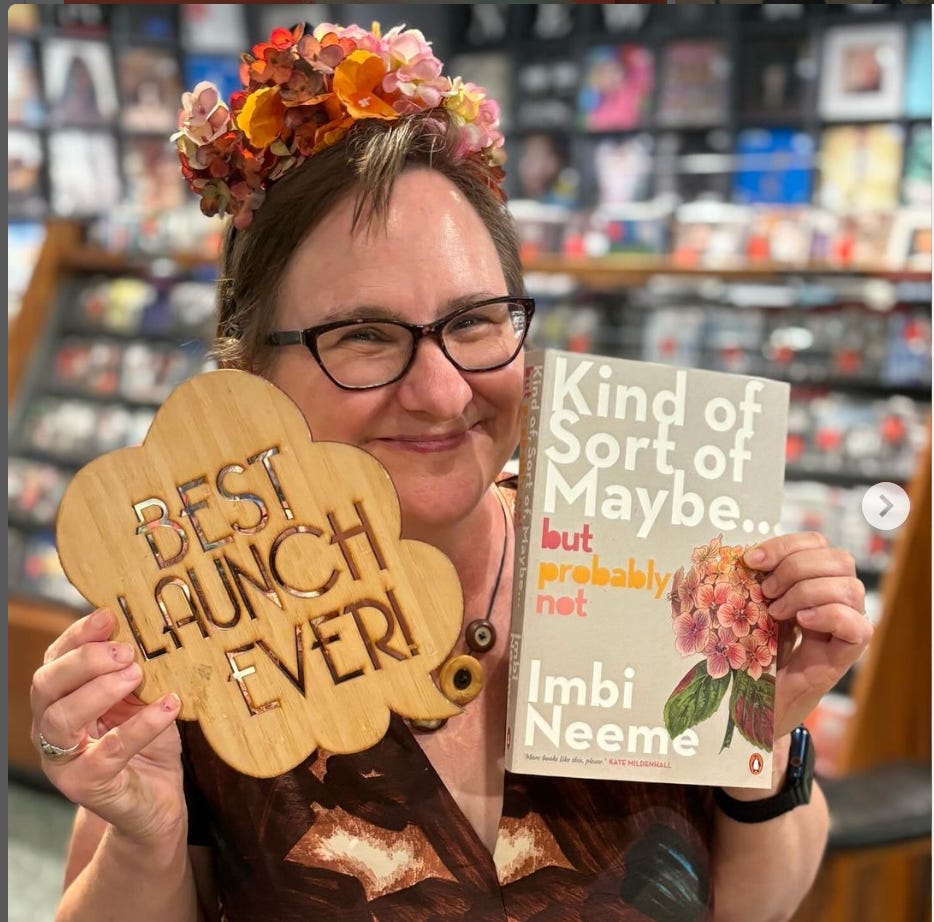The same Queen in a hydrangea crown holding up a book and a cheap-looking plywood sign in the shape of a speech bubble that says "BEST LAUNCH EVER". 