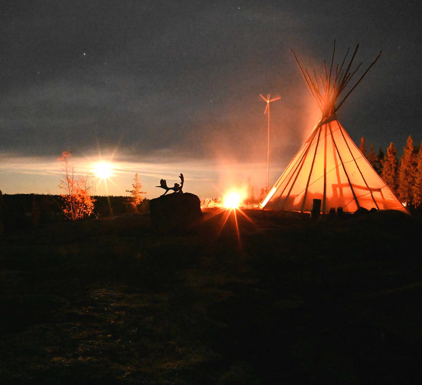 The moon shines brightly near a tipi. There is a fire inside and just outside the tipi. Antlers sit on top of a rock in the middle.