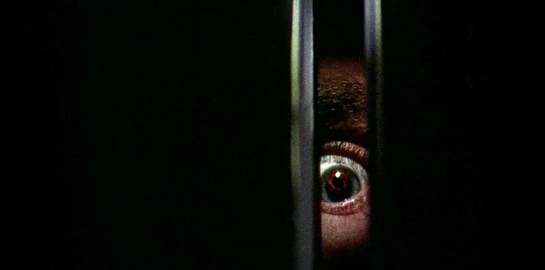 A close-up of a wide, bloodshot eye peering through a narrow crack in the darkness, evoking a sense of dread and suspense. This is a haunting still from the 1974 horror film Black Christmas.