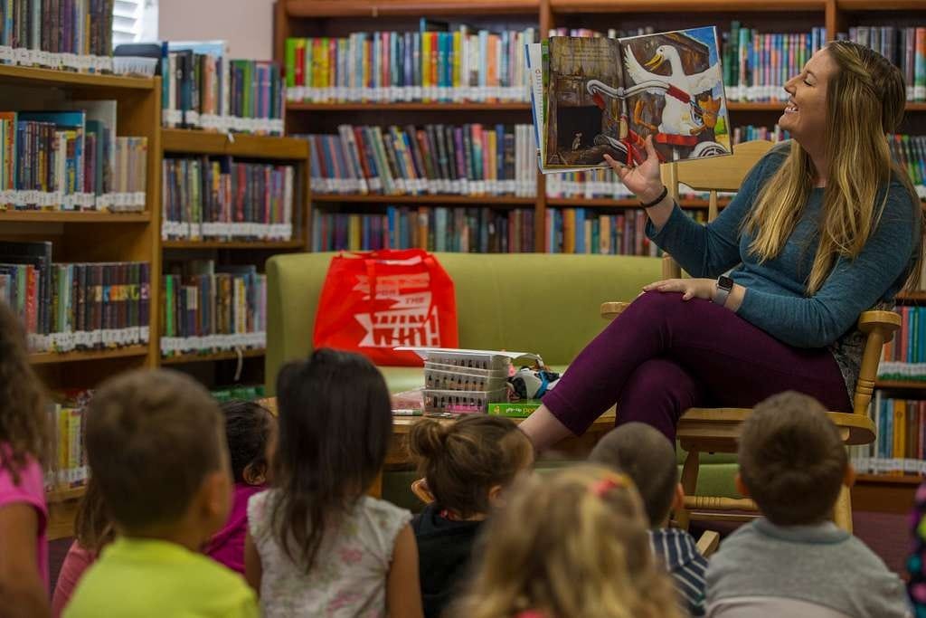 A librarian sitting in a wooden rocking chair smiles and holds up a picture book as she reads to a group of young children in a well-lit library with inviting bookshelves surrounding them 