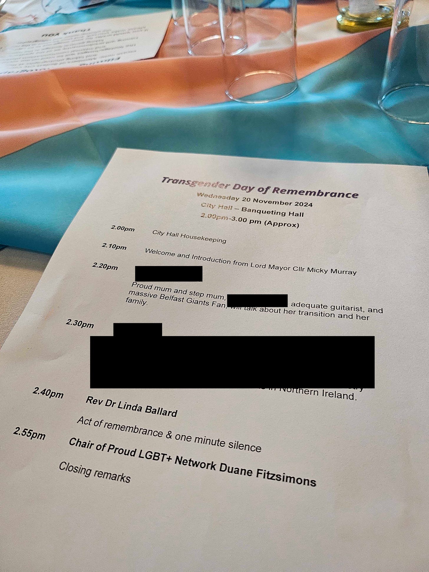 A programme sheet for the Transgender Day of Remembrance event on Wednesday, 20 November 2024, at City Hall, Banqueting Hall. The sheet lists the schedule from 2.00pm to 3.00pm, including City Hall housekeeping, a welcome from Lord Mayor Cllr Micky Murray, and various speakers. Sections of the text have been redacted. The event includes speeches, an act of remembrance, a minute of silence, and closing remarks. The sheet is placed on a table with the transgender pride flag colours (blue, pink, and white) as part of the decor, alongside glassware.