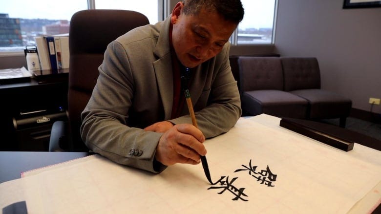 A man in blazer writes on a piece of paper with ink brush inside an office.