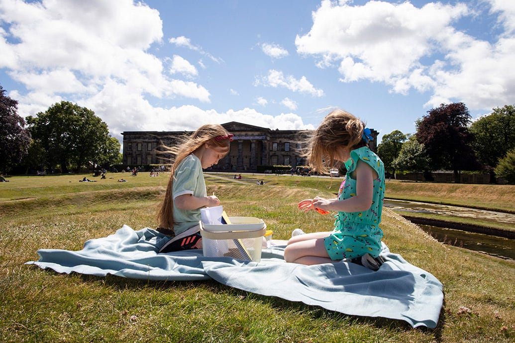 Two girls sitting on a picnic blanket on a sunny day. They are playing with items from the art basket with the Modern One building in the background.