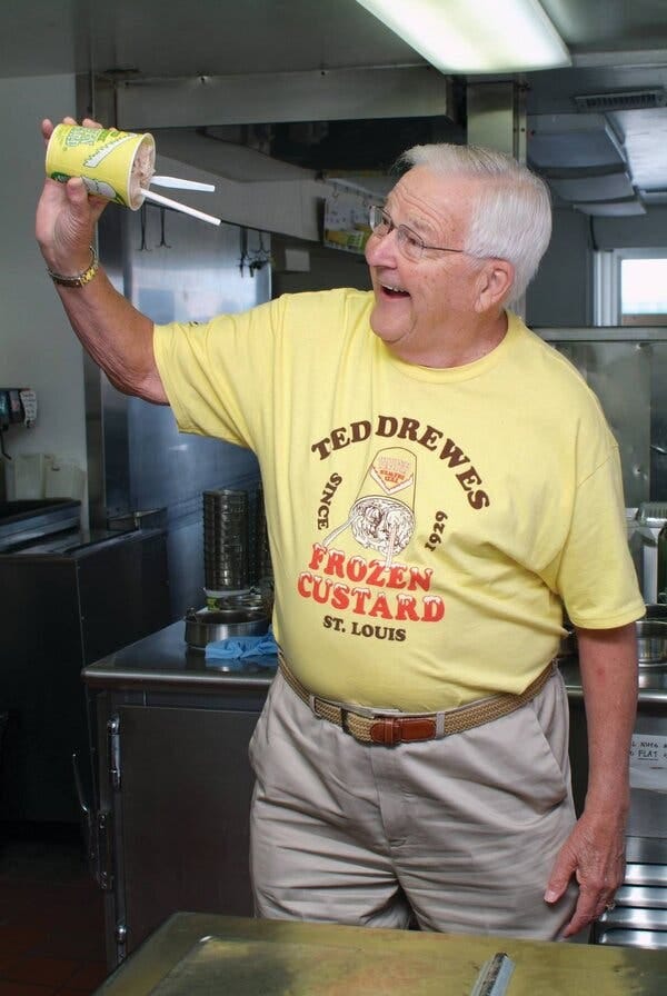 Ted Drewes Jr., a white-haired man with glasses wearing a yellow “Ted Drewes Frozen Custard” T-shirt, smiles as he holds a cup of frozen custard horizontally.