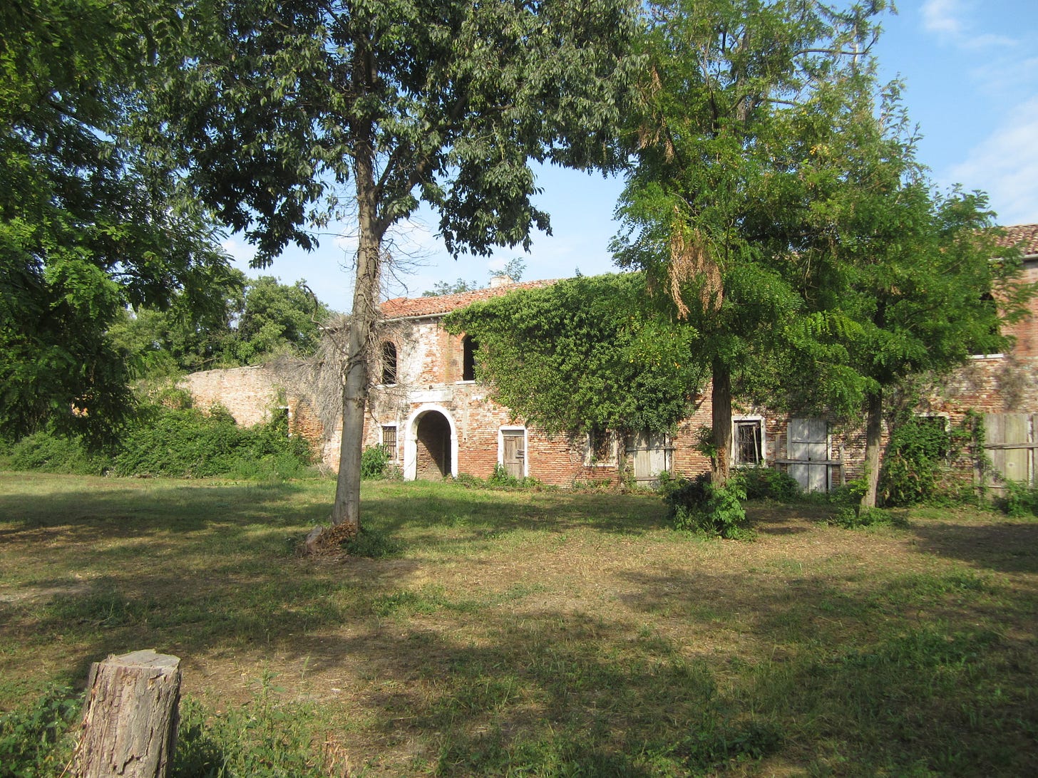 A modern photo of a dilapidated two-storey brick building with an arched doorway. Behind it is a clear sky and in front there is grass and trees casting shadows, making the scene appear tranquil.