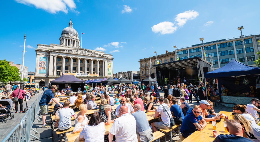 Nottingham's Old Market Square with people sat at rows of wooden benches, with purple and blue stalls in front of them