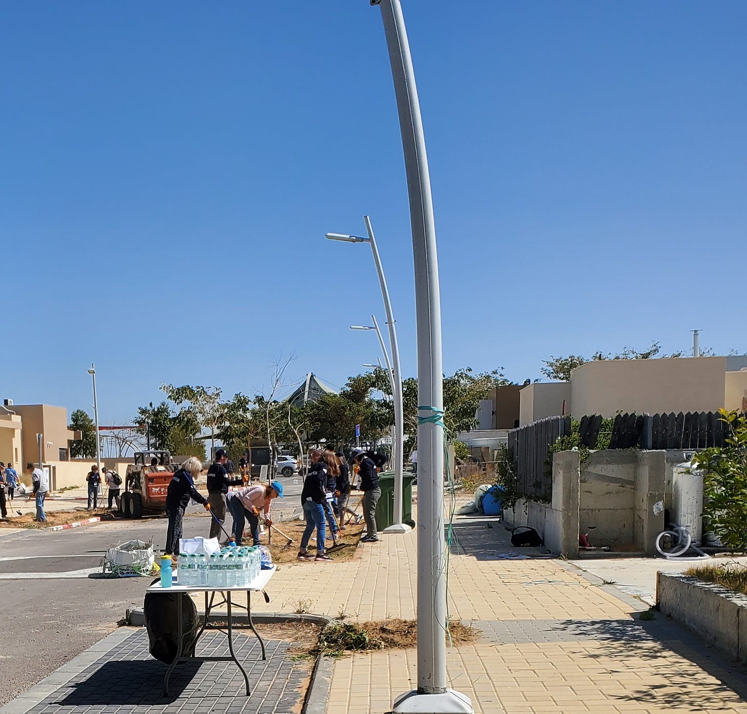 A group of volunteers with garden tools in front of square, concrete homes under a blue cloudless sky. 