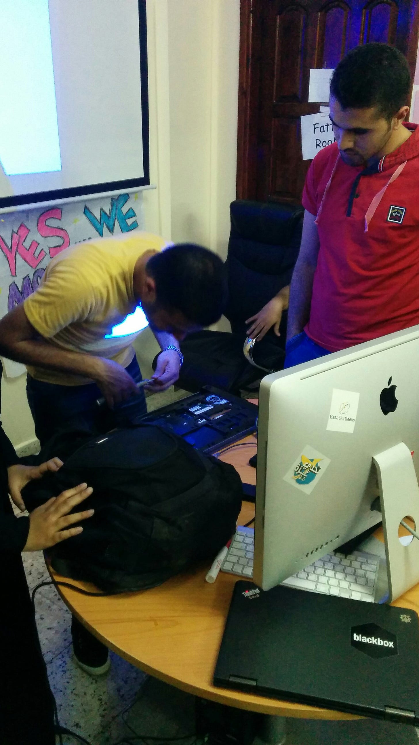 A man hunches over a laptop working to repair it, while a projector screen displays behind him