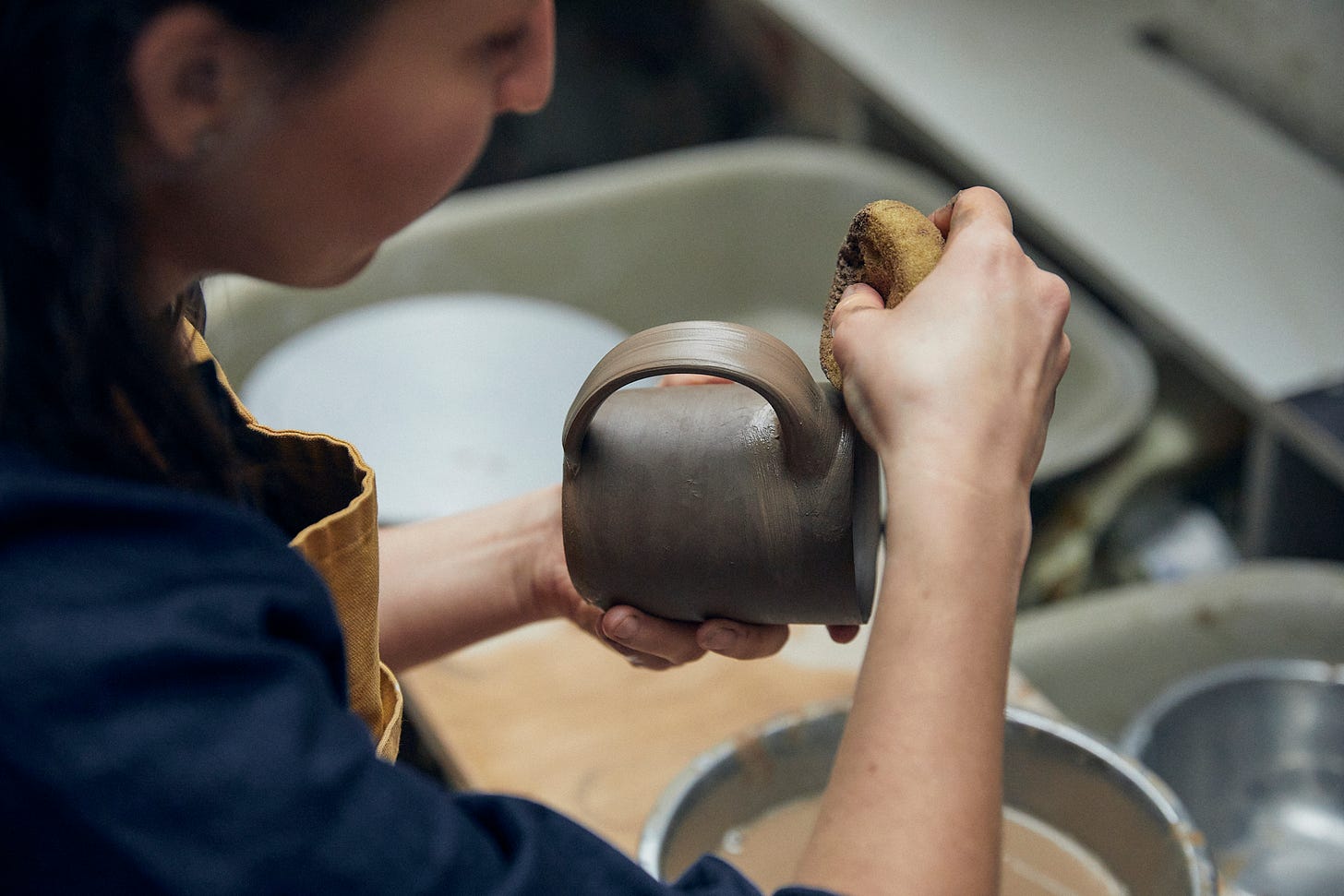 Seeing over the shoulder of a female ceramicist as she works. She is using a sponge to smooth the joint of a handle on a mug.