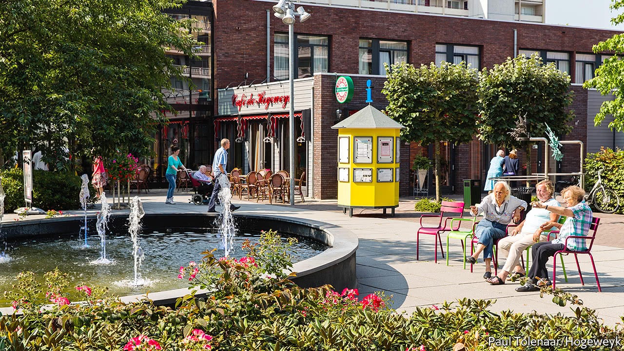 The open-air plaza in the dementia village, with trees and shrubs, large fountain, and tables and chairs for sitting among the cafe, grocery store, and other spaces.