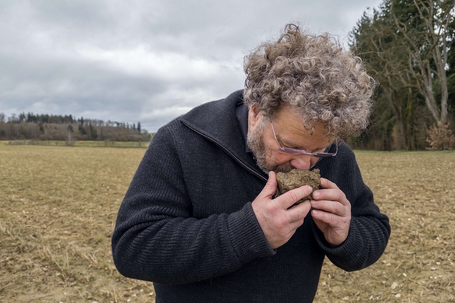 Der Fachmann für regenerative Landwirtschaft Axel Vohwinkel, ein Mann mit lockigem Haar und Brille, hält ein Stück Erde vor seine Nase und riecht daran, um die Bodenqualität zu beurteilen. Im Hintergrund erstreckt sich ein Feld unter einem bewölkten Himmel. Der Fachmann für regenerative Landwirtschaft Axel Vohwinkel, ein Mann mit lockigem Haar und Brille, hält ein Stück Erde vor seine Nase und riecht daran, um die Bodenqualität zu beurteilen. Im Hintergrund erstreckt sich ein Feld unter einem bewölkten Himmel.