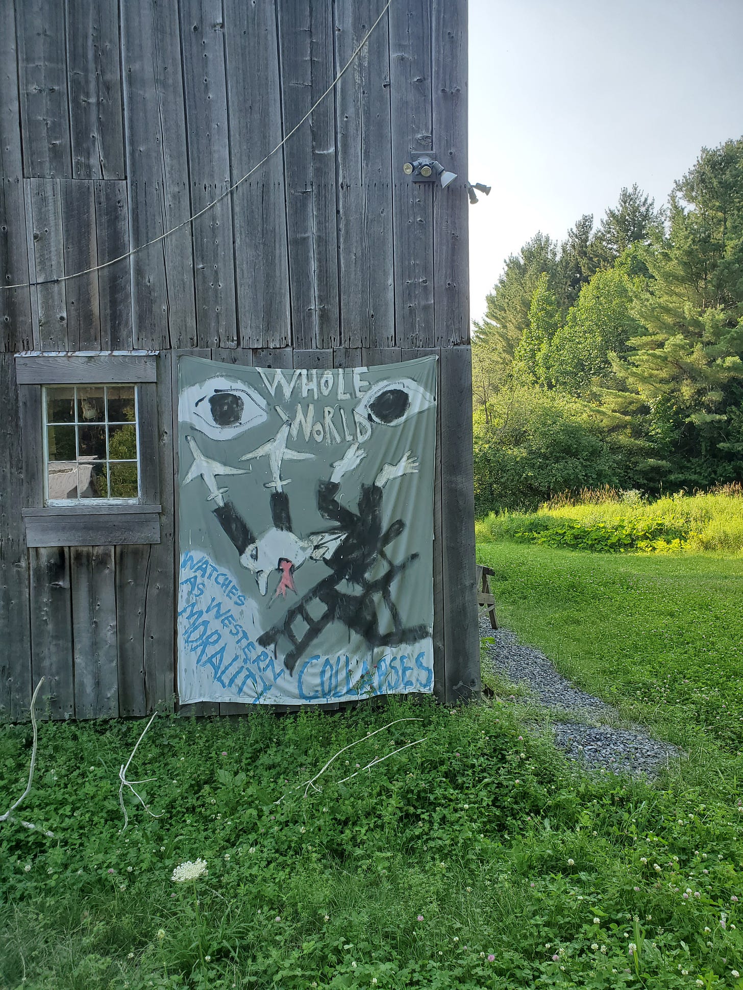 A handpainted banner on the side of a gray barn depicts large eyes and a human figure in a suit, falling off a chair, its limbs turning into birds and planes. Text surrounding reads "Whole world watches as Western morality collapses".