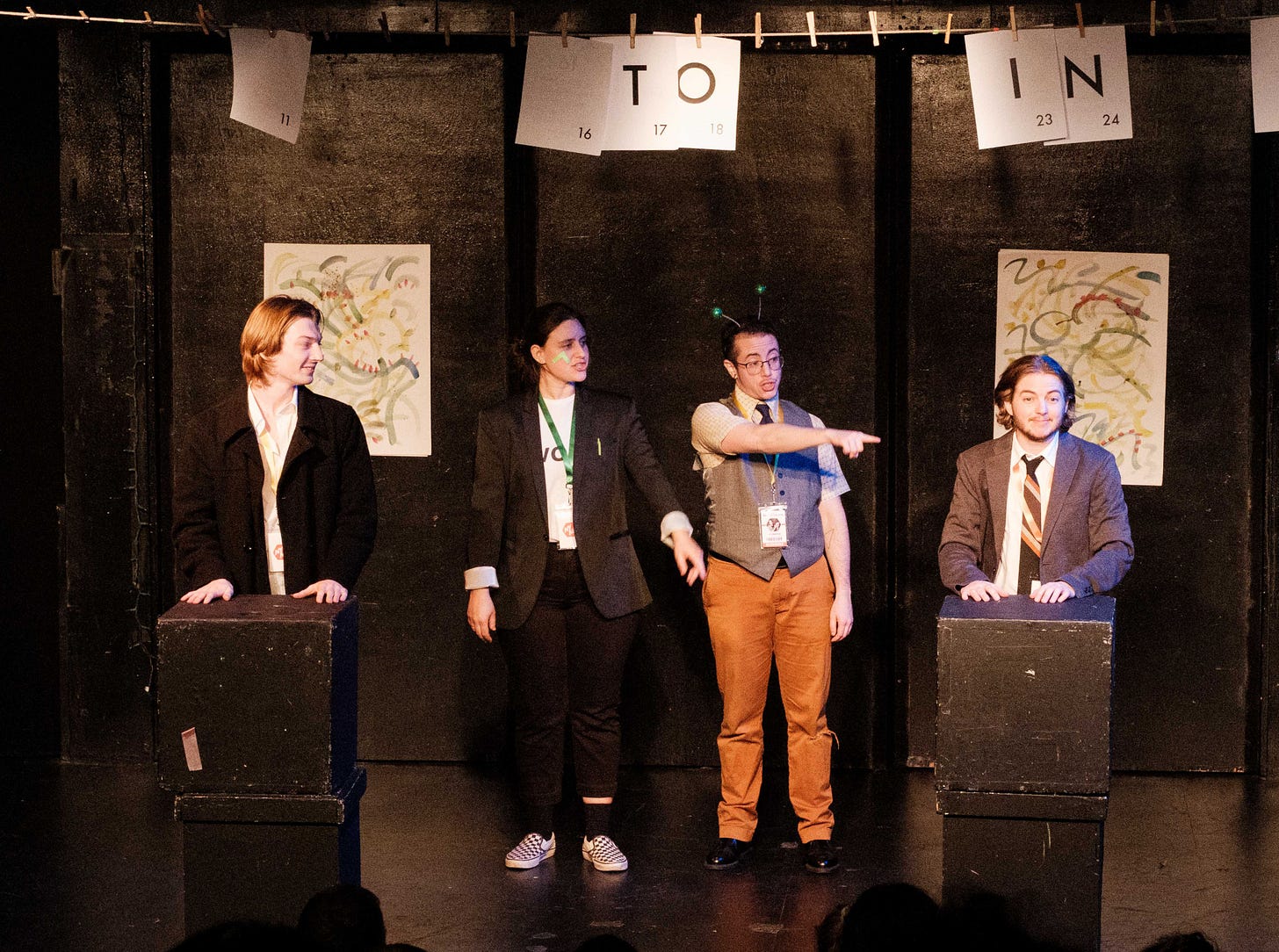 Four white performers stand on a black stage. Three of them are looking and pointing at Jack, on the far right, who is looking to the audience with a shrug. All wear business attire. Jack and a performer on the left stand behind two stacked blocks each. Four white performers stand on a black stage. Three of them are looking and pointing at Jack, on the far right, who is looking to the audience with a shrug. All wear business attire. Jack and a performer on the left stand behind two stacked blocks each.