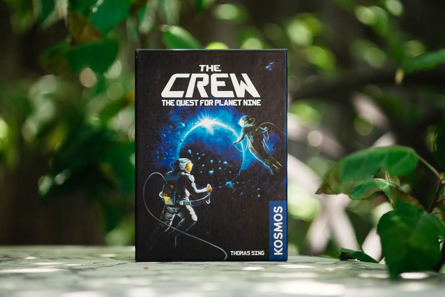 The box for the card game The Crew: The Quest for Planet Nine displayed on a marble table with green leaves in the background.