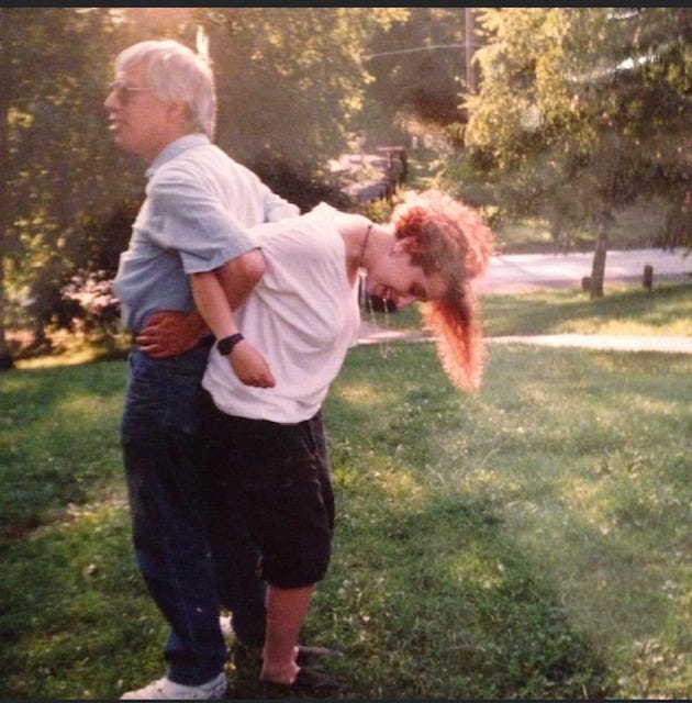 Gray haired olive skinned man links arms, back to back, with laughing, pale redheaded teenage girl