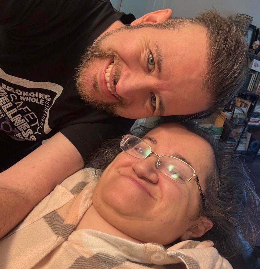 Photo of Stefan and Athena in their living room. Athena is reclined in her wheelchair and Stefan is leaning over her to take the photo.