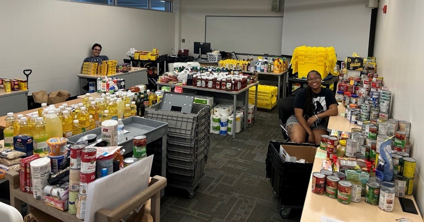 A room filled with organized food donation, including canned goods, condiments and cooking oil, with two people seated among the items, organizing the donations. A room filled with organized food donation, including canned goods, condiments and cooking oil, with two people seated among the items, organizing the donations.
