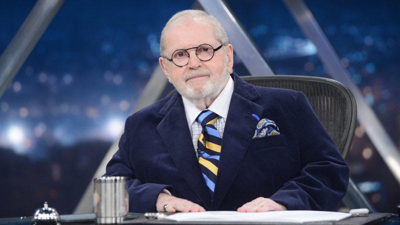 Imagem do Jô Soares sentado à mesa de seu programa na Globo. Está de terno escuro e gravata listrada em tons de azul e amarelo.