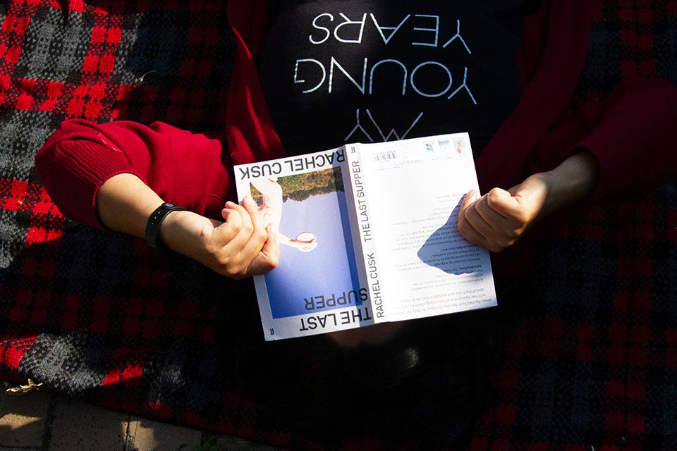 A person reading a copy of Rachel Cusk's 'The Last Supper' on a red tartan picnic blanket.