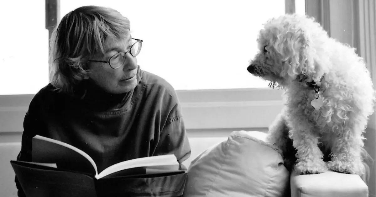 A black and white photo of Mary Oliver who is sitting on a couch with a book in her hand. She is looking to her left at her dog who is sitting on the arm of the couch.