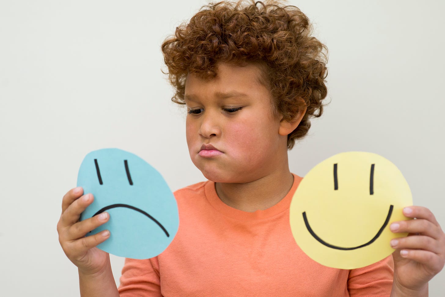 A visual of a child holding circular shaped emojis. One hand holding yellow smily emoji, the other hand holding a blue coloured sad emoji.