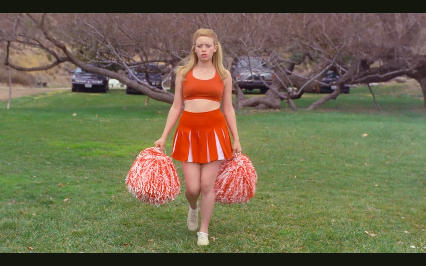 A young Natasha Lyonne in her role as the main character of "But I'm a Cheerleader" walking in cheerleading costume with pom poms over grass, cars behind a tree in the background, she looks sad and determined