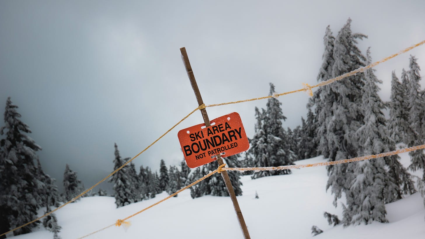 A snowy scene with trees depicts a ski resort with a red sign that reads Ski Area Boundary Not Patrolled. The sign is red and held up by a pole. The trees are evergreens