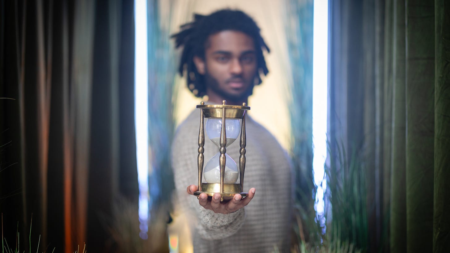 A dark-skinned man with dreadlocks holds out a giant egg timer towards the viewer.
