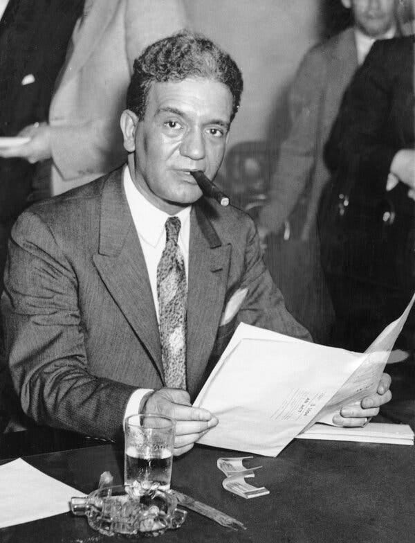 A black-and-white photograph of a suited man with a cigar in his mouth sitting at a table. He is looking up from an unfolded document that he holds in his hands. An ashtray and a glass of water sit on the table near his right hand.
