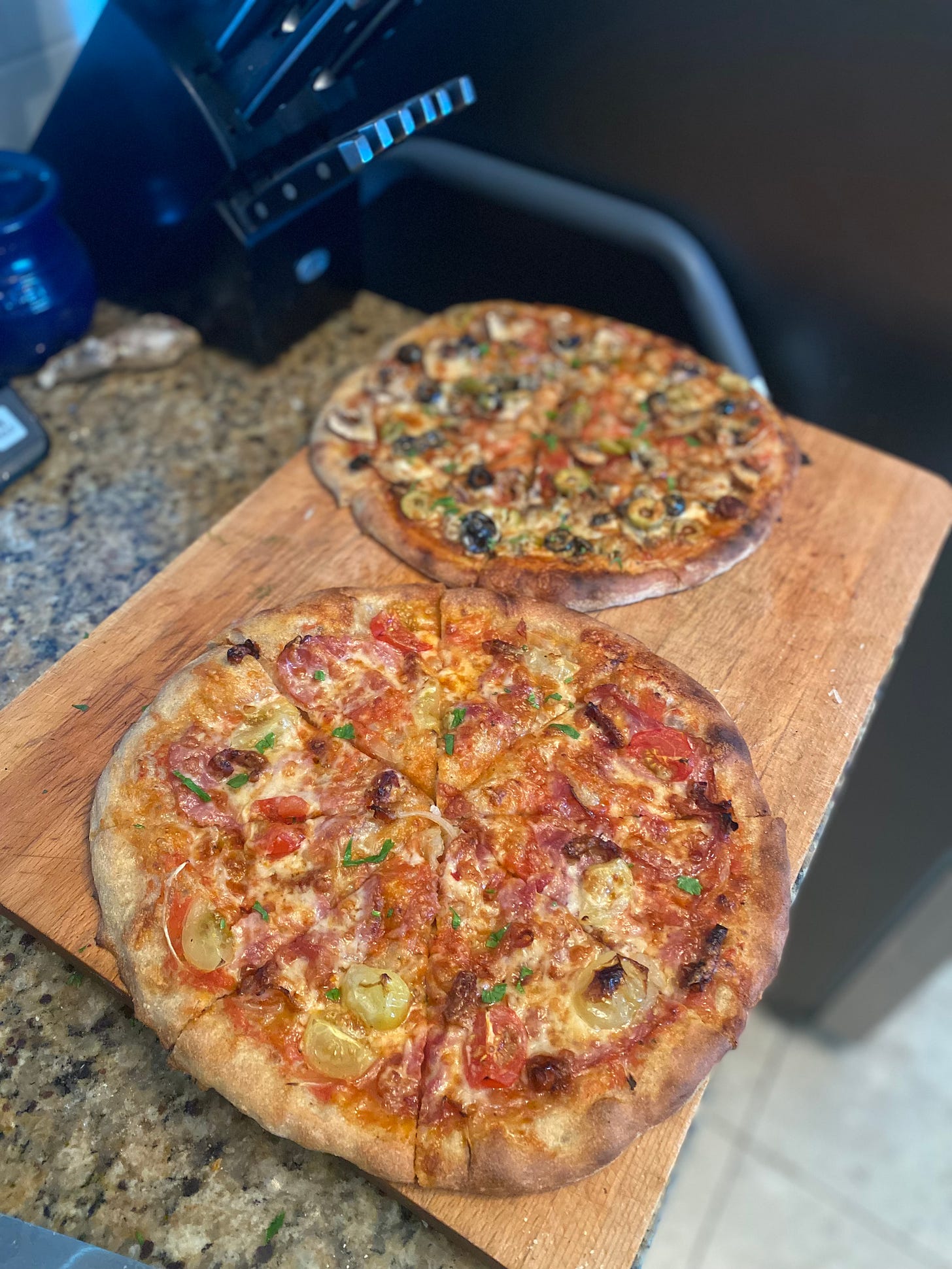 The two pizzas described above, the olive one in the the background and the tomato one in the foreground. The tomato pieces are red, orange, and yellow. 