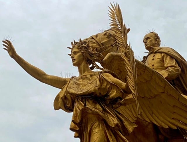 A golden statue of a winged Victory, her right arm extended, in front of General Sherman riding his horse, Ontario. She clutches a leafy frond in her hand.