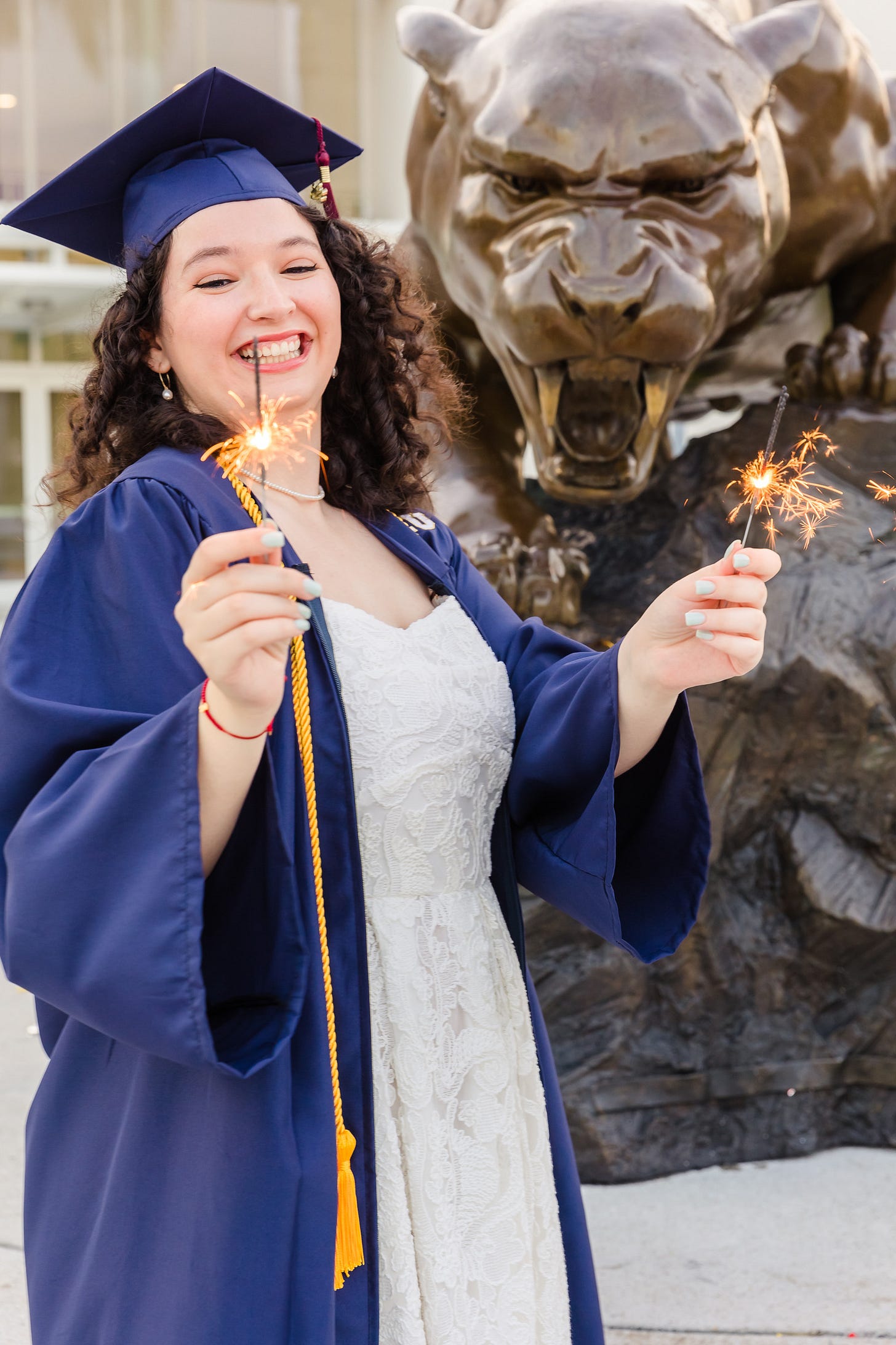 Photo of a young woman wear a white dress beneath an opened graduation gown, with graduation cap, and holding lit firework sparklers, with a bronze statue of a panther behind her.