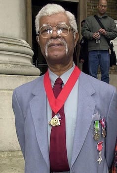 An older man dressed in a pale blue suit and red tie with his MBE award around his neck.