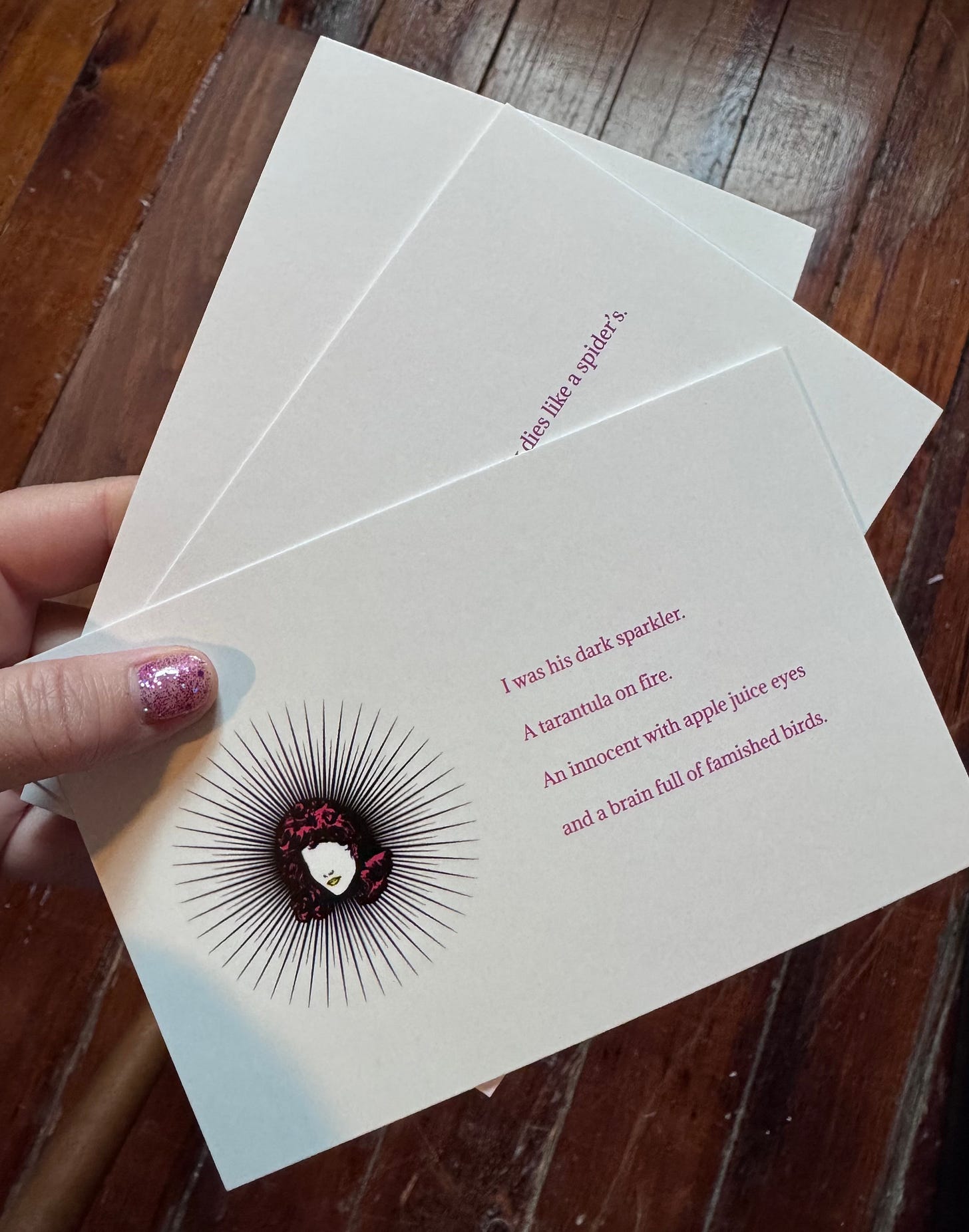 The front of a postcard. It matches the book cover for "Dark Sparkler" with a white background and a drawing of the head of a faceless woman with bright pink hair. Around the head are lines shooting out so that she looks like she's in the middle of a starburst. To the right, pink text reads "I was his dark sparkler. A tarantula on fire. An innocent with apple juice eyes and a brain full of famished birds."
