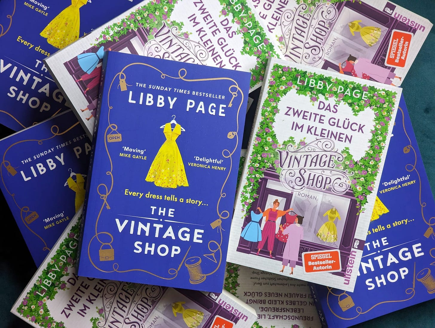 A pile of copies of The Vintage Shop in English and in German. The English version has a bright blue cover with a yellow dress in the middle. The German version shows a shopfront surrounded by flowers, with a yellow dress in the window and two figures looking at dresses.