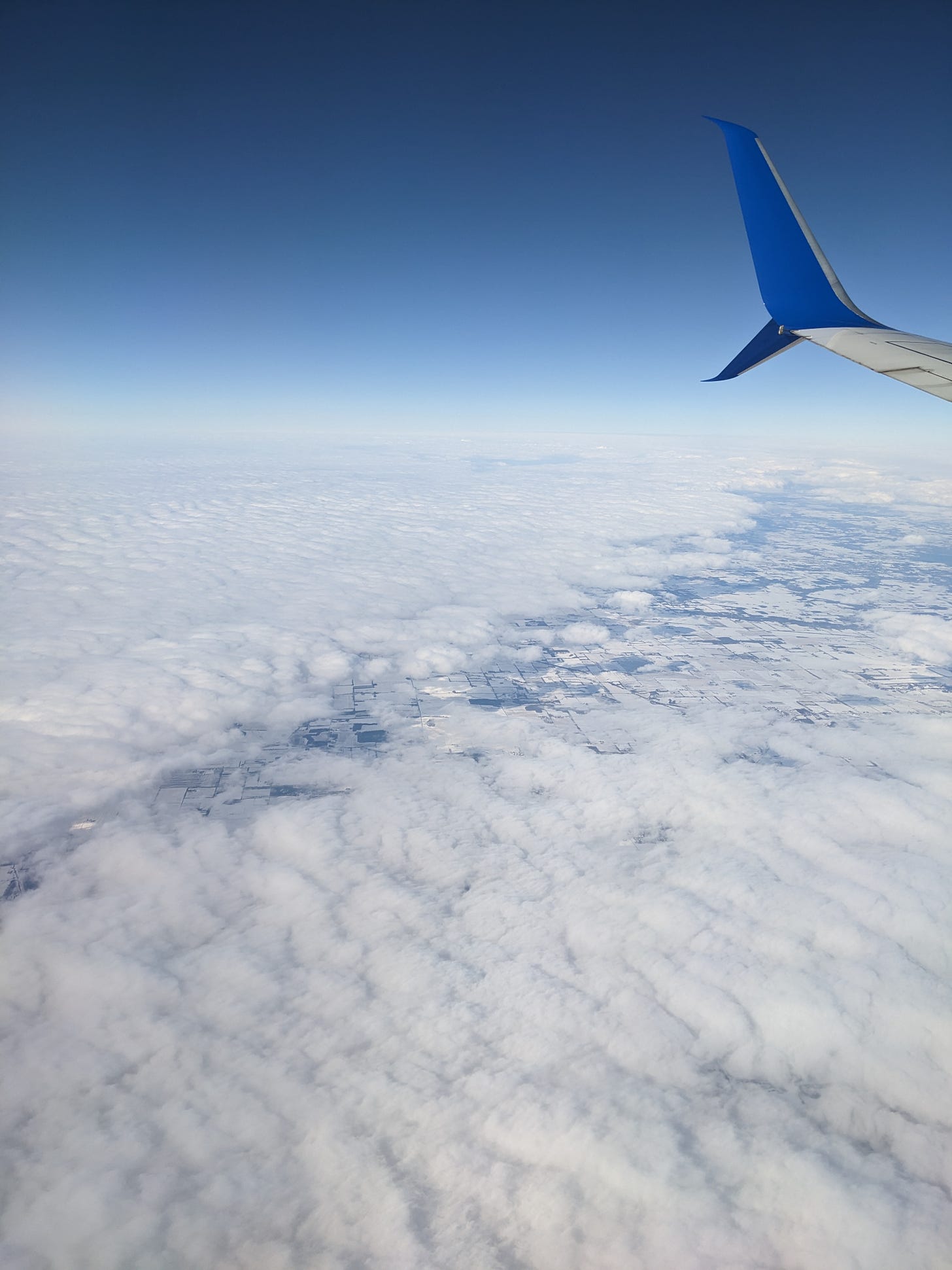 blue sky and a bit of ground breaking through the clouds, with the edge of an airplane wing on the right side