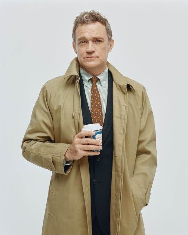 Mr. Keefe poses in a khaki-colored trench coat over a navy suit jacket, pinstriped shirt and tan print tie. He holds a takeout paper cup. 