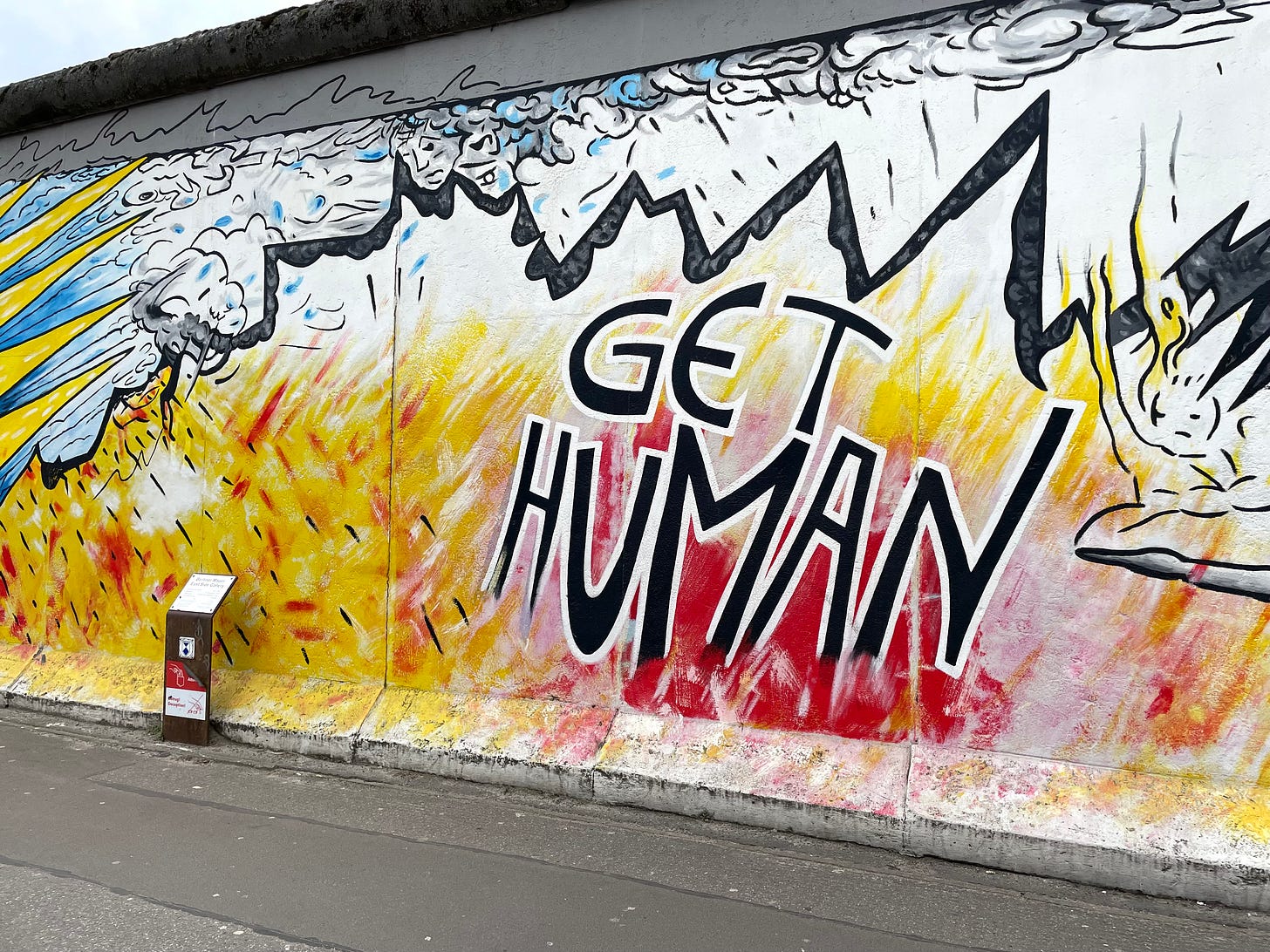 A vibrant mural painted on a section of the Berlin Wall, featuring bold text 'GET HUMAN' surrounded by jagged black lines, fiery yellow and red flames, and stormy clouds. A small informational plaque stands nearby, adding historical context to the artwork.