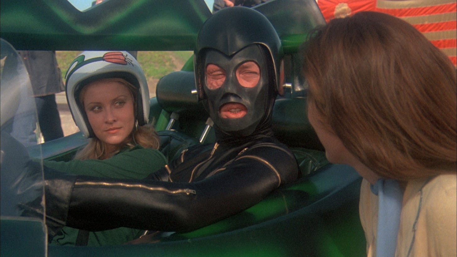 Movie still from Death Race 2000. A man covered in leather and a leather mask sits in a car, a woman in his passenger seat and another woman talking to them from outside.