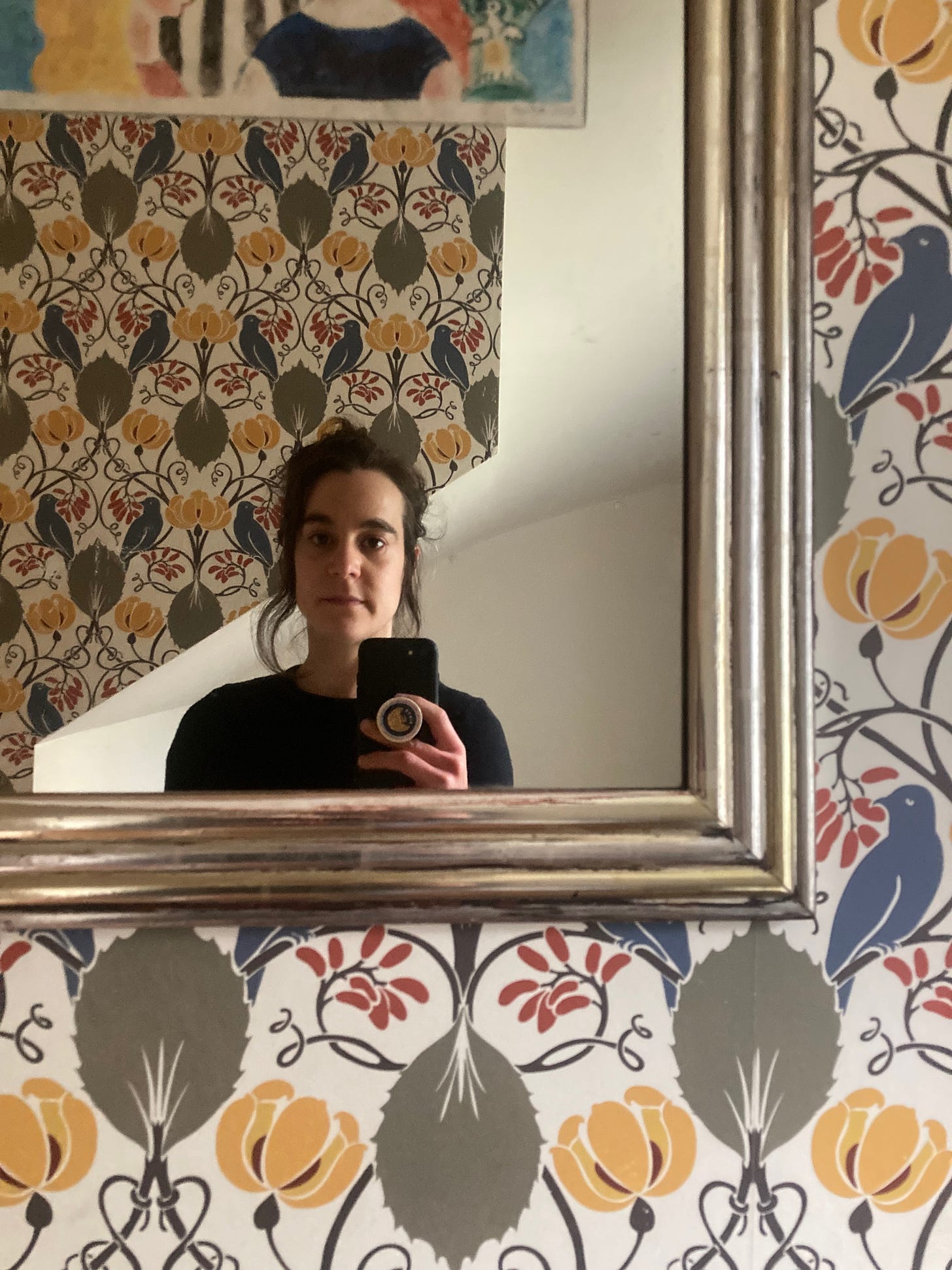 A selfie of siloh looking in a mirror on a wall with antique wallpaper, art deco bird pattern