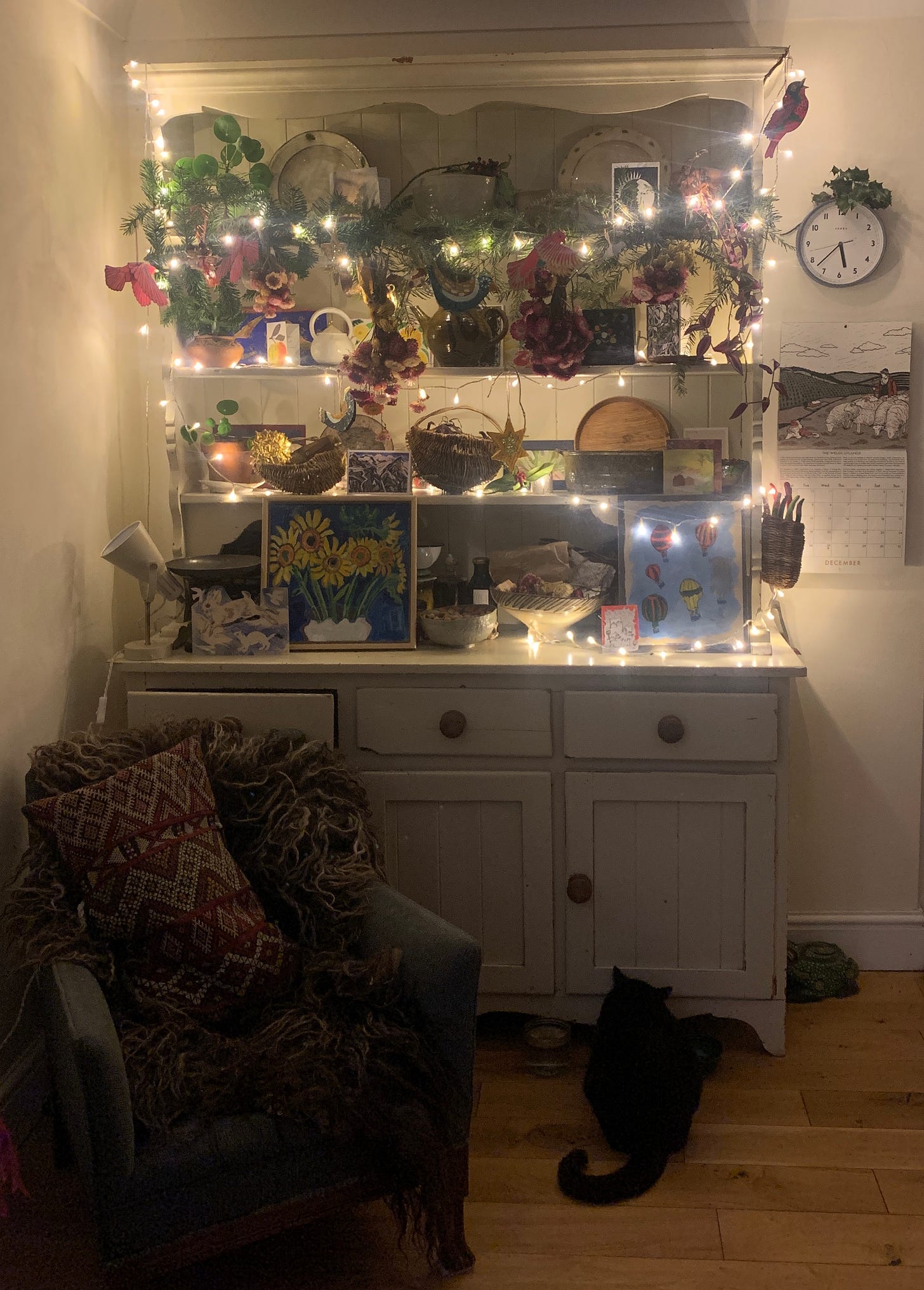 A white kitchen dresser filled with knick knacks, fairy lights, evergreen branches, pictures, woven baskets. A black cat sits at the bottom